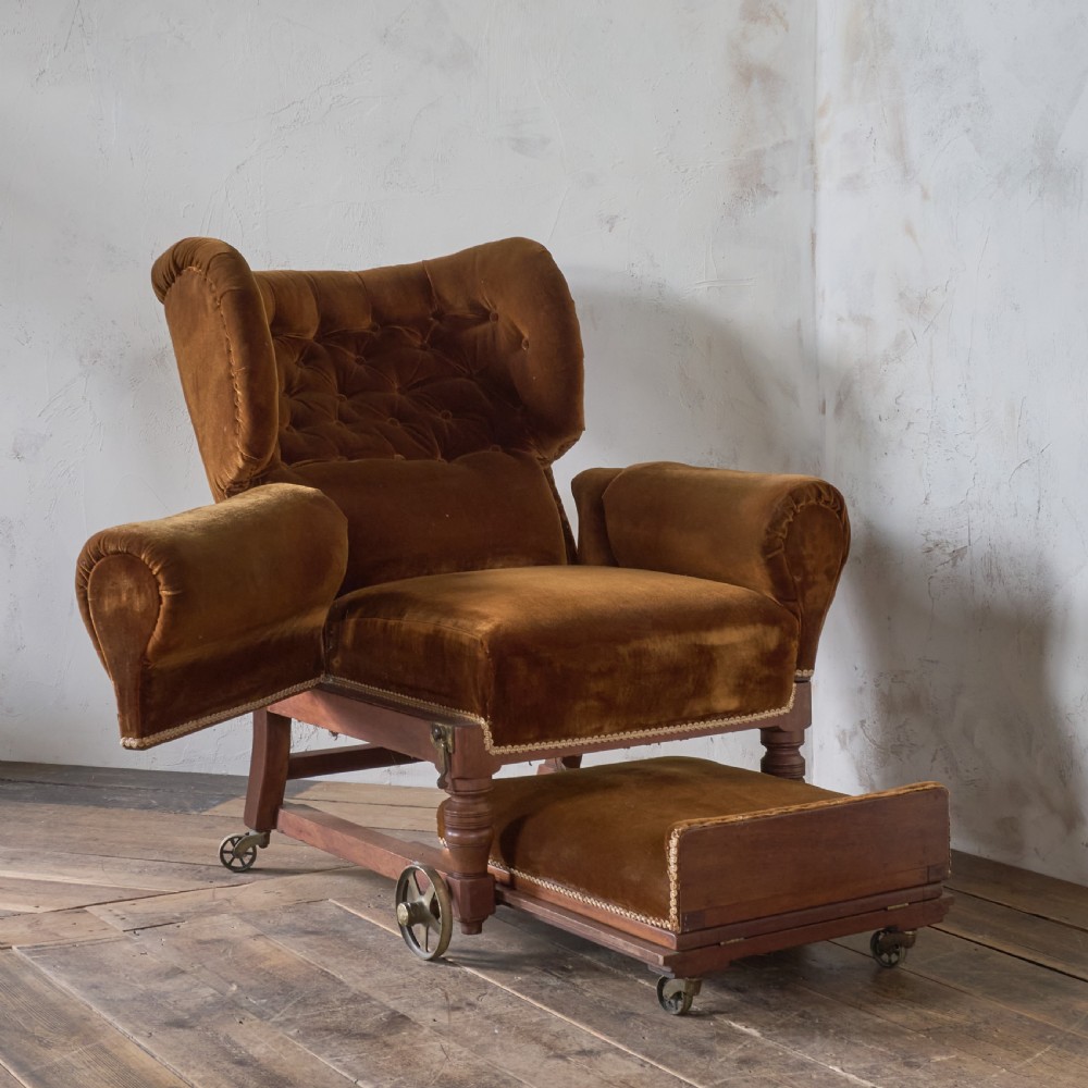 An antique Victorian mechanical reclining armchair upholstered in brown velvet with a button-tufted backrest, padded armrests, and an extendable footrest on brass castors.