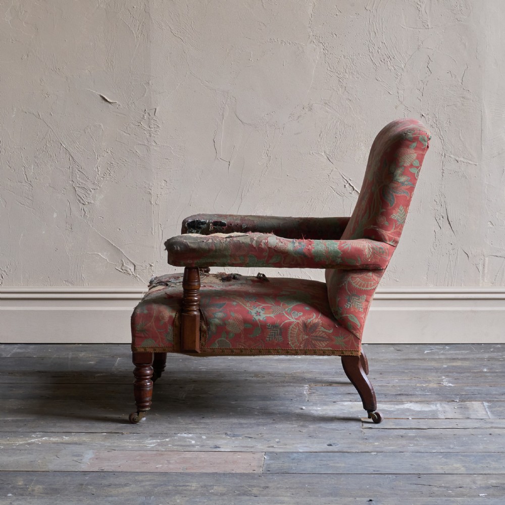 A vintage open armchair featuring a wooden frame, turned front legs on castors, and worn patterned pink upholstery. View X