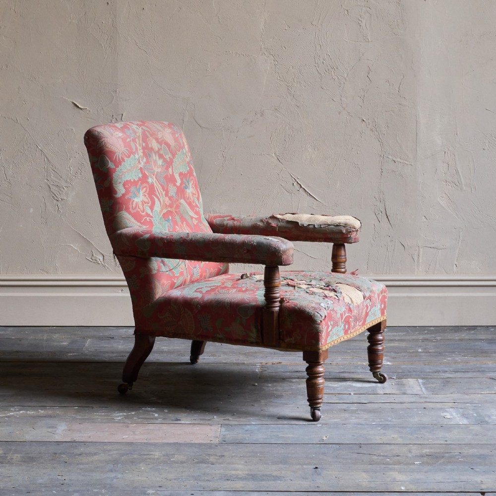 A vintage open armchair featuring a wooden frame, turned front legs on castors, and worn patterned pink upholstery.