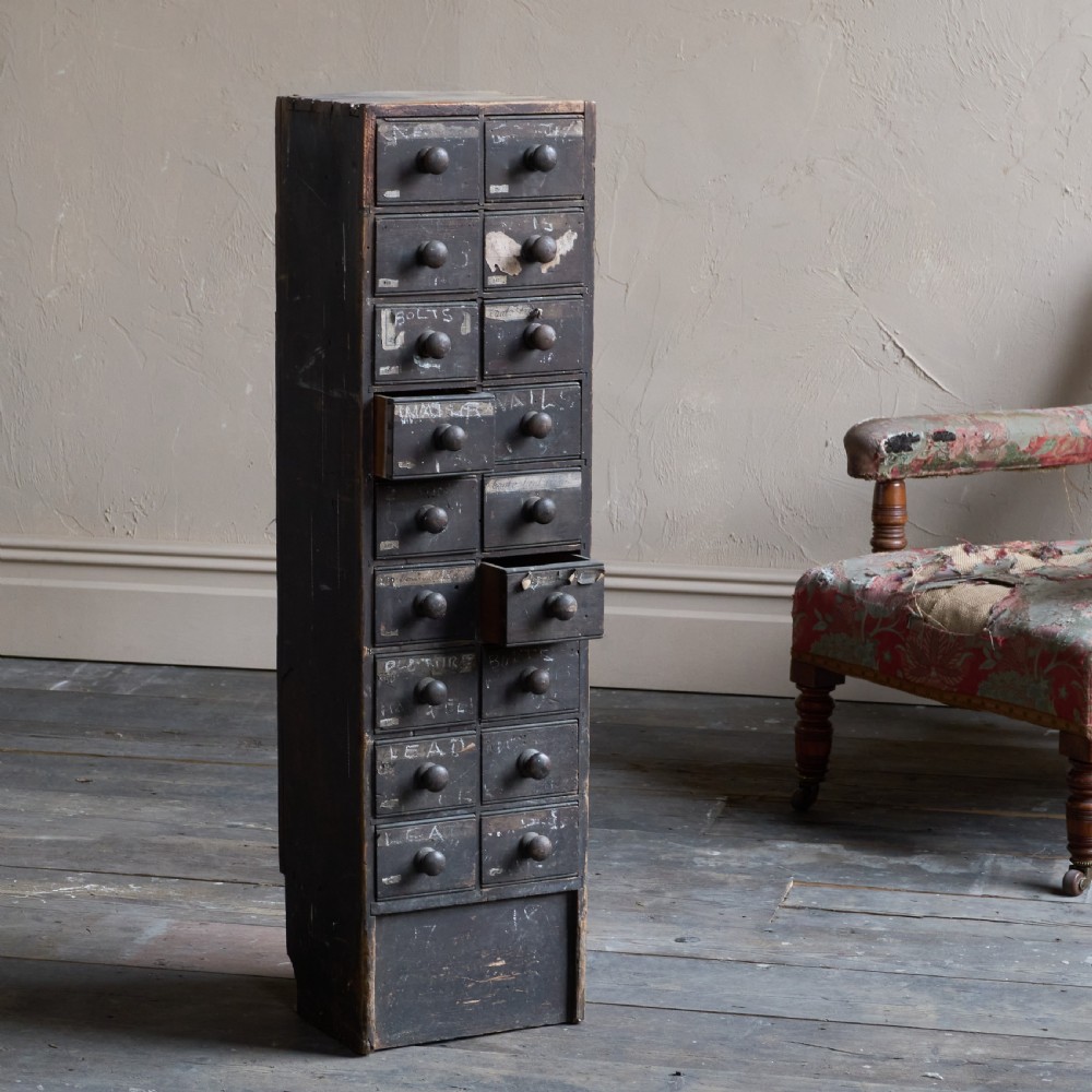 A tall and narrow antique dark wood ironmongers chest featuring sixteen small drawers with turned knobs and hand-painted white labels.