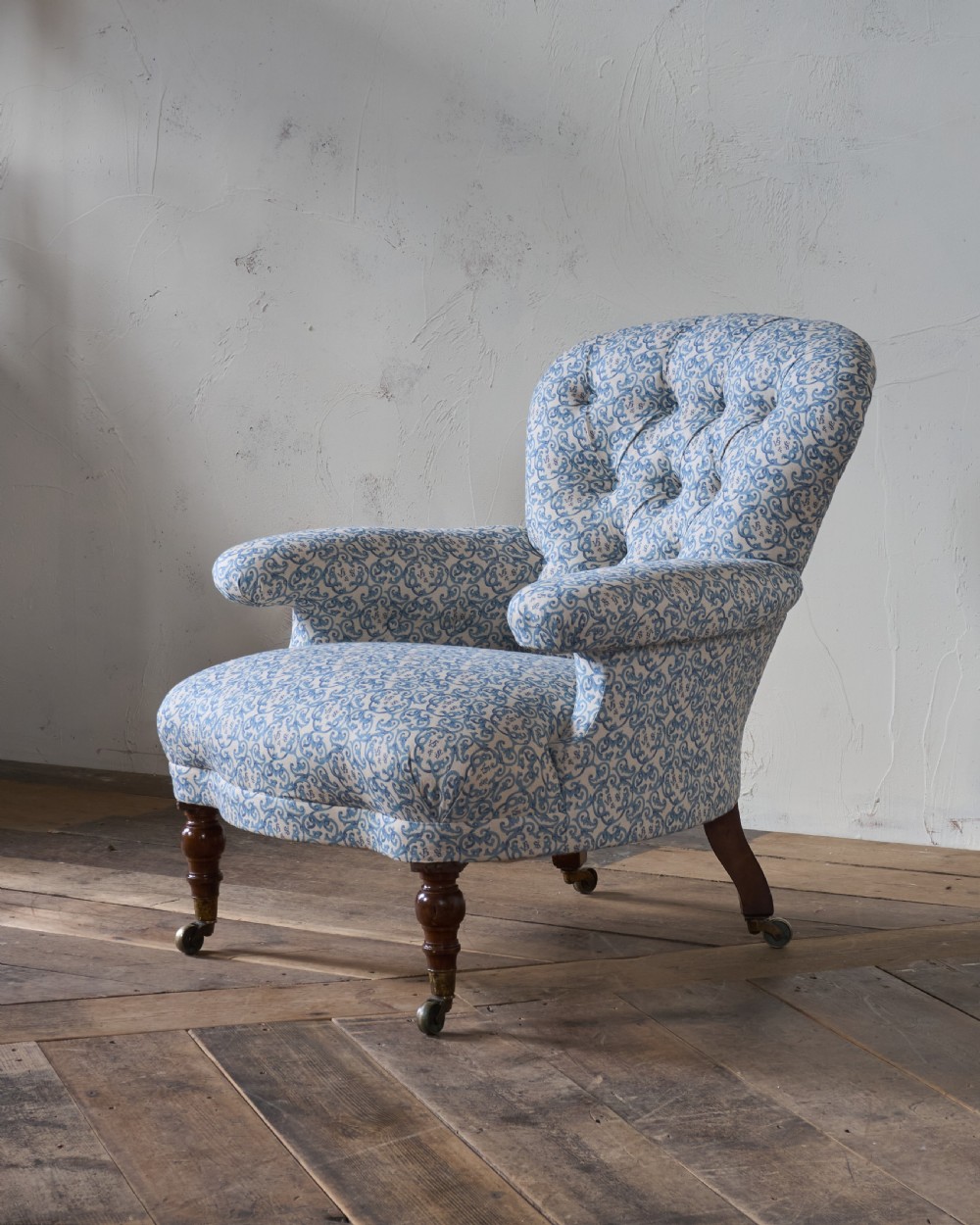 A Victorian Howard style armchair upholstered in blue and white scroll printed fabric with deep buttoning and turned mahogany legs on brass castors.