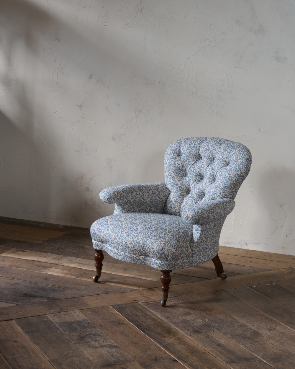 A Victorian Howard style armchair upholstered in blue and white scroll printed fabric with deep buttoning and turned mahogany legs on brass castors. View VI
