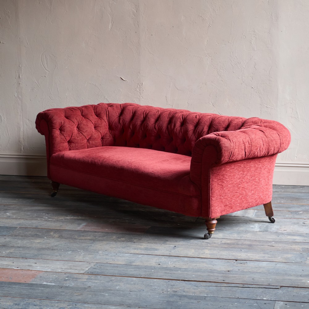 A deep red fabric Chesterfield sofa featuring all-over button-tufted upholstery, rolled arms, and turned wooden legs on brass castors.