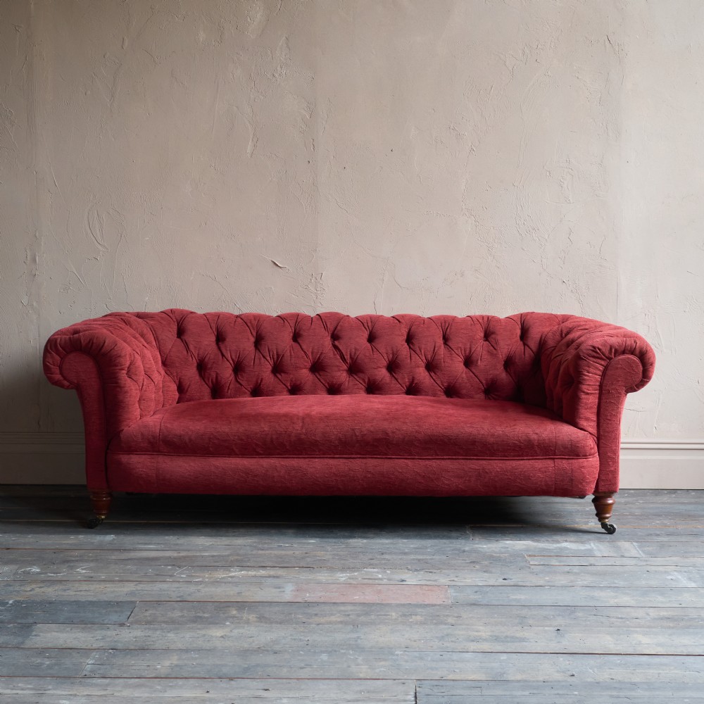 A deep red fabric Chesterfield sofa featuring all-over button-tufted upholstery, rolled arms, and turned wooden legs on brass castors. View II