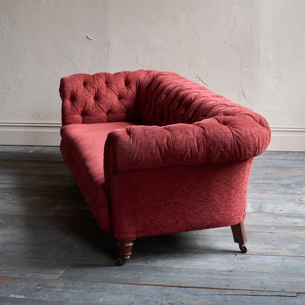 A deep red fabric Chesterfield sofa featuring all-over button-tufted upholstery, rolled arms, and turned wooden legs on brass castors. View III