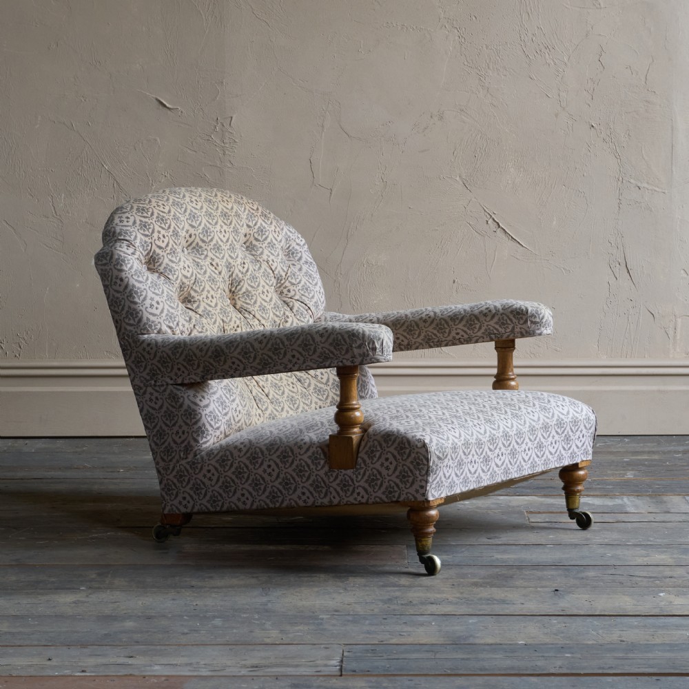 An antique open armchair featuring patterned grey ticking upholstery, a button-tufted backrest, padded armrests on turned wooden uprights, and short turned legs with brass castors.