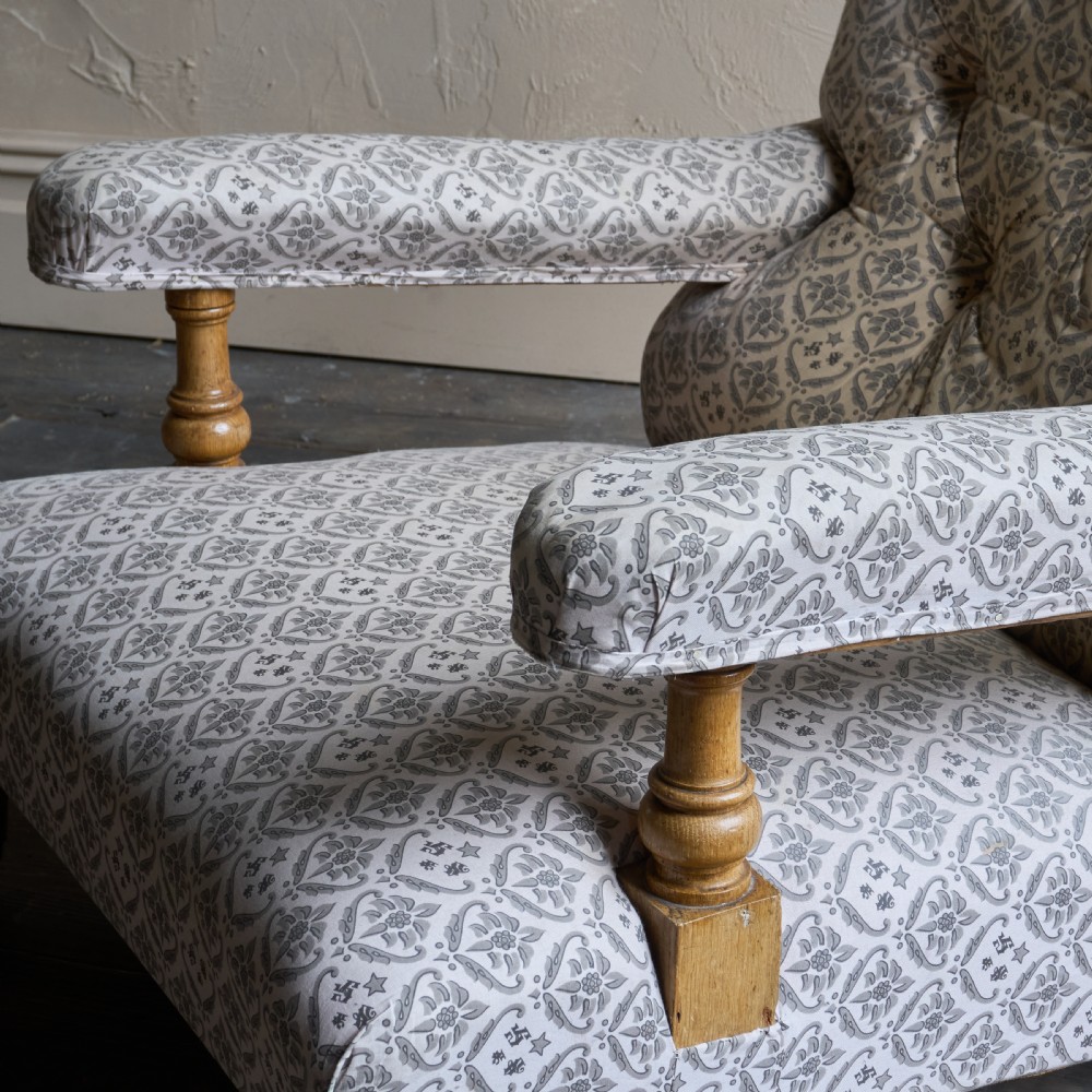 An antique open armchair featuring patterned grey ticking upholstery, a button-tufted backrest, padded armrests on turned wooden uprights, and short turned legs with brass castors. View V