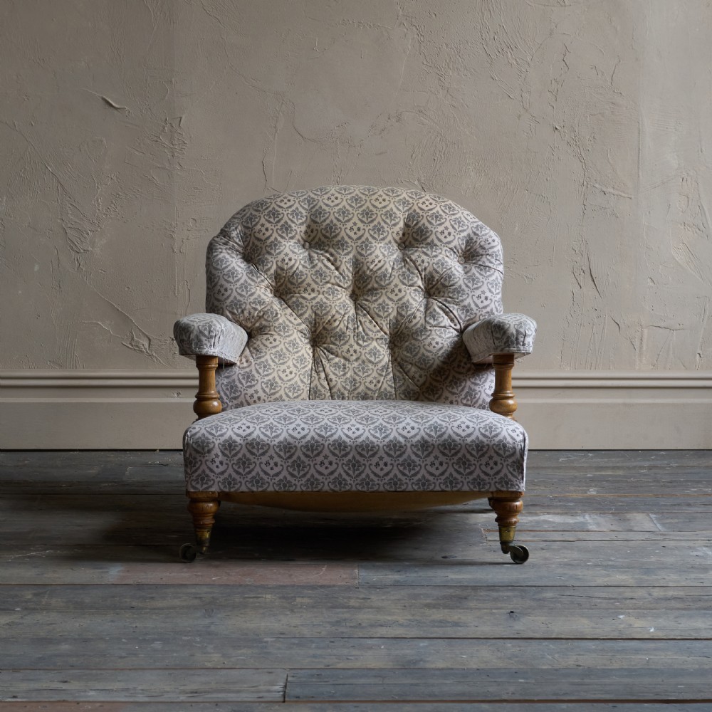 An antique open armchair featuring patterned grey ticking upholstery, a button-tufted backrest, padded armrests on turned wooden uprights, and short turned legs with brass castors. View XII