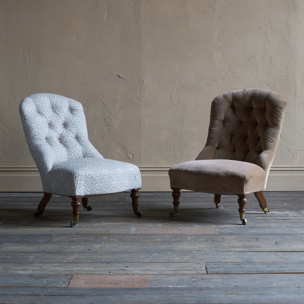 A pair of Victorian low-slung button-backed slipper chairs with turned wooden legs on brass castors, one in taupe velvet and one in patterned blue and white fabric.