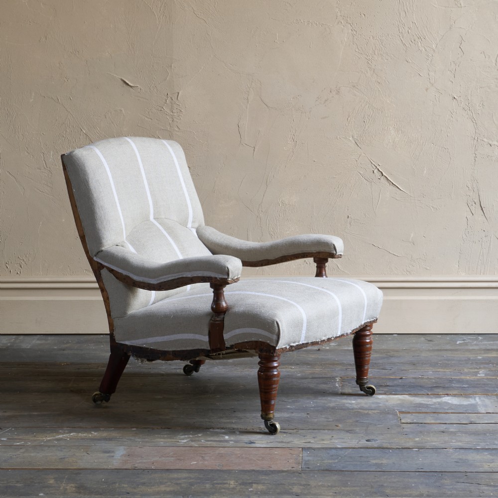 An antique open armchair upholstered in cream fabric with vertical white ticking stripes, featuring turned wooden front legs with brass castors.