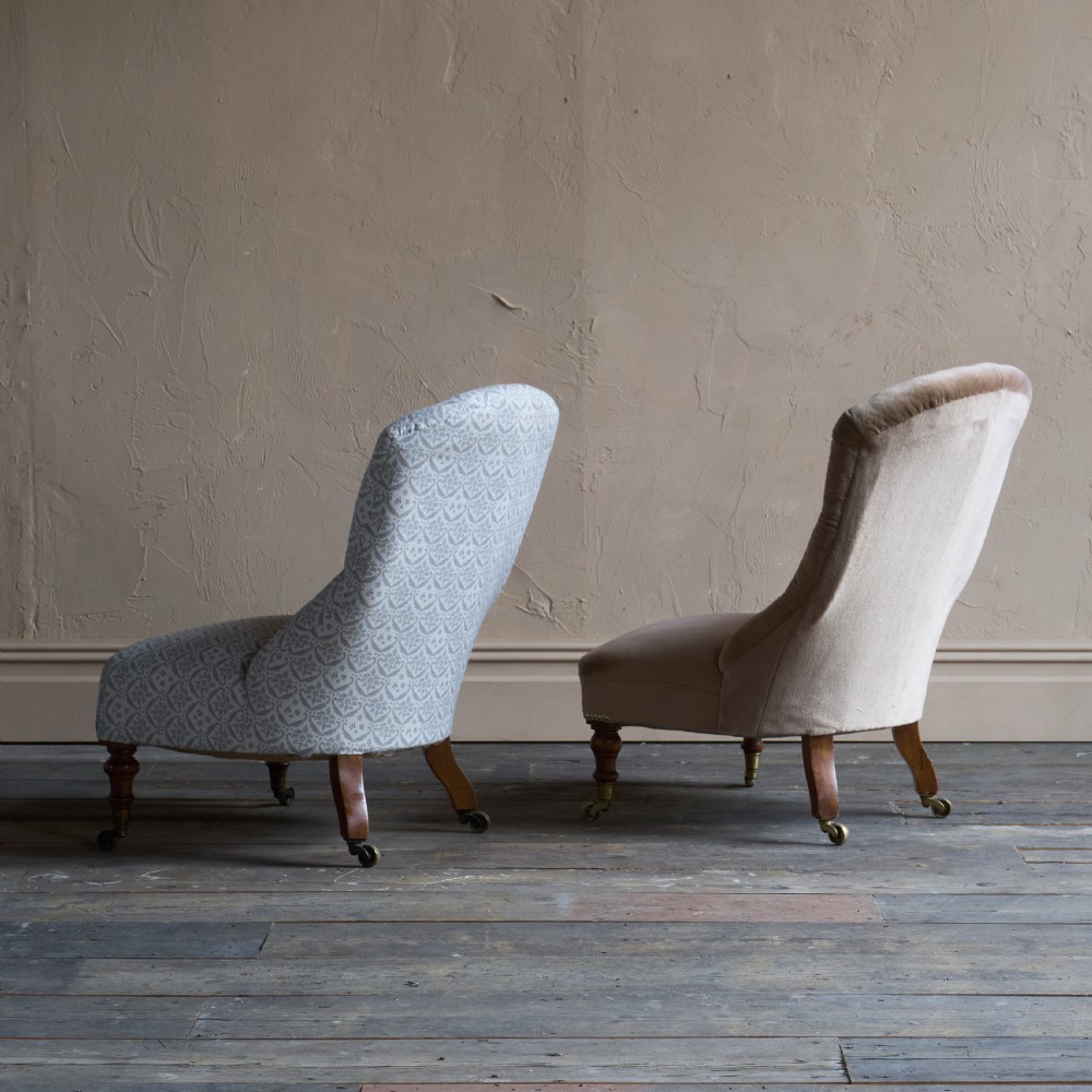 A pair of Victorian low-slung button-backed slipper chairs with turned wooden legs on brass castors, one in taupe velvet and one in patterned blue and white fabric. View X