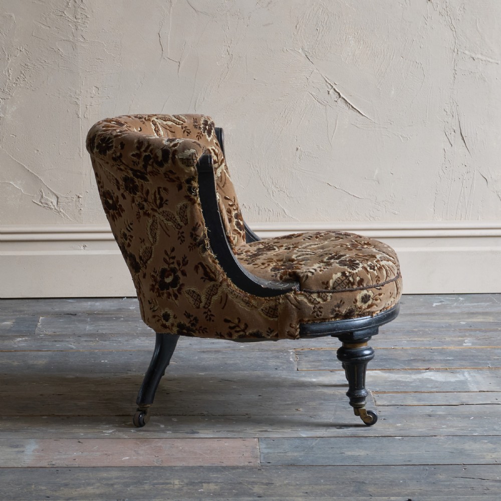 A Victorian low-seated tub chair upholstered in brown patterned floral velvet with ebonised turned legs, gilt accents, and brass castors. View V