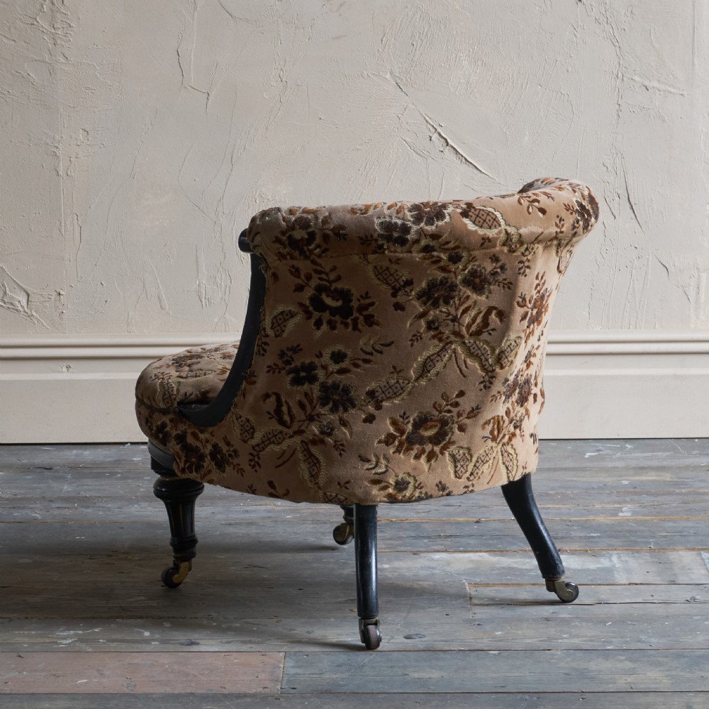 A Victorian low-seated tub chair upholstered in brown patterned floral velvet with ebonised turned legs, gilt accents, and brass castors. View VI