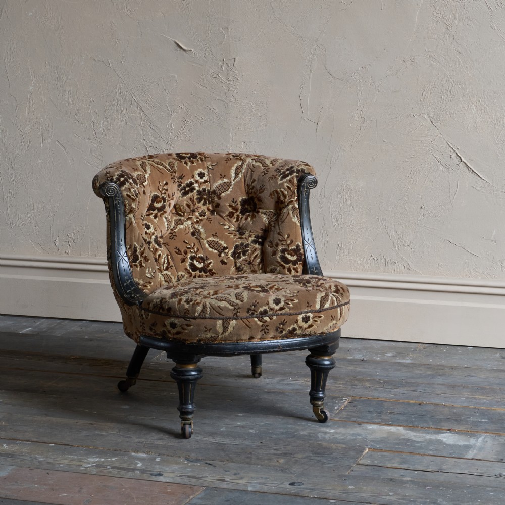 A Victorian low-seated tub chair upholstered in brown patterned floral velvet with ebonised turned legs, gilt accents, and brass castors. View VII