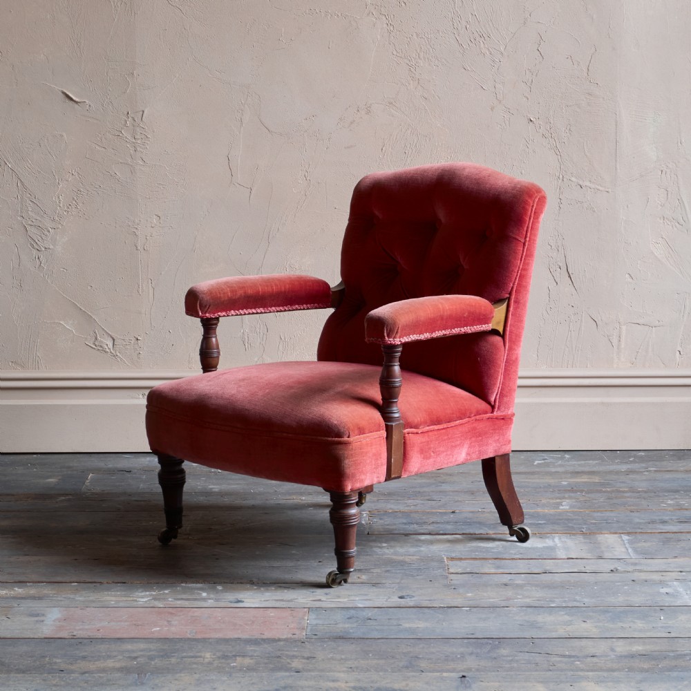 Victorian open armchair featuring deep buttoned rose pink velvet upholstery, turned wooden front legs, and brass castors.