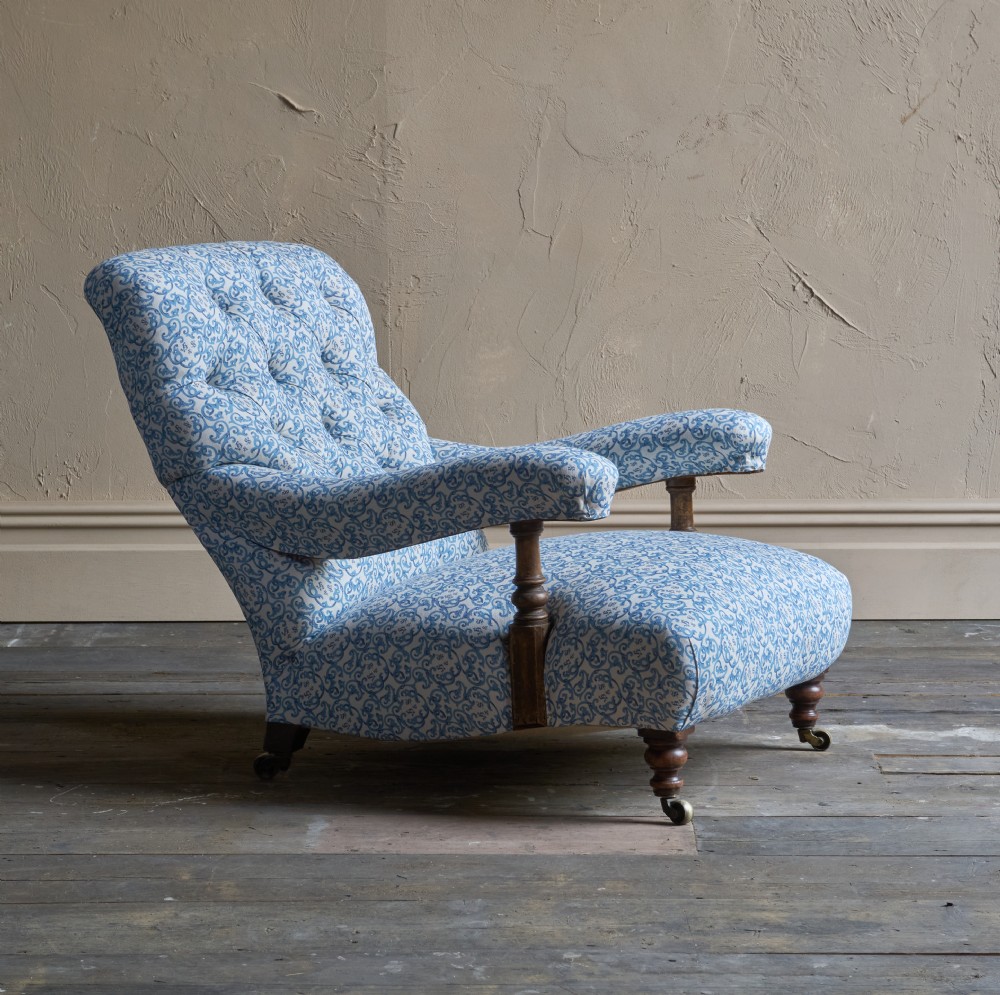 A low Victorian open armchair upholstered in a blue and white scroll pattern, featuring deep buttoning on the backrest, padded arms on turned wooden supports, and turned legs on castors. View VI