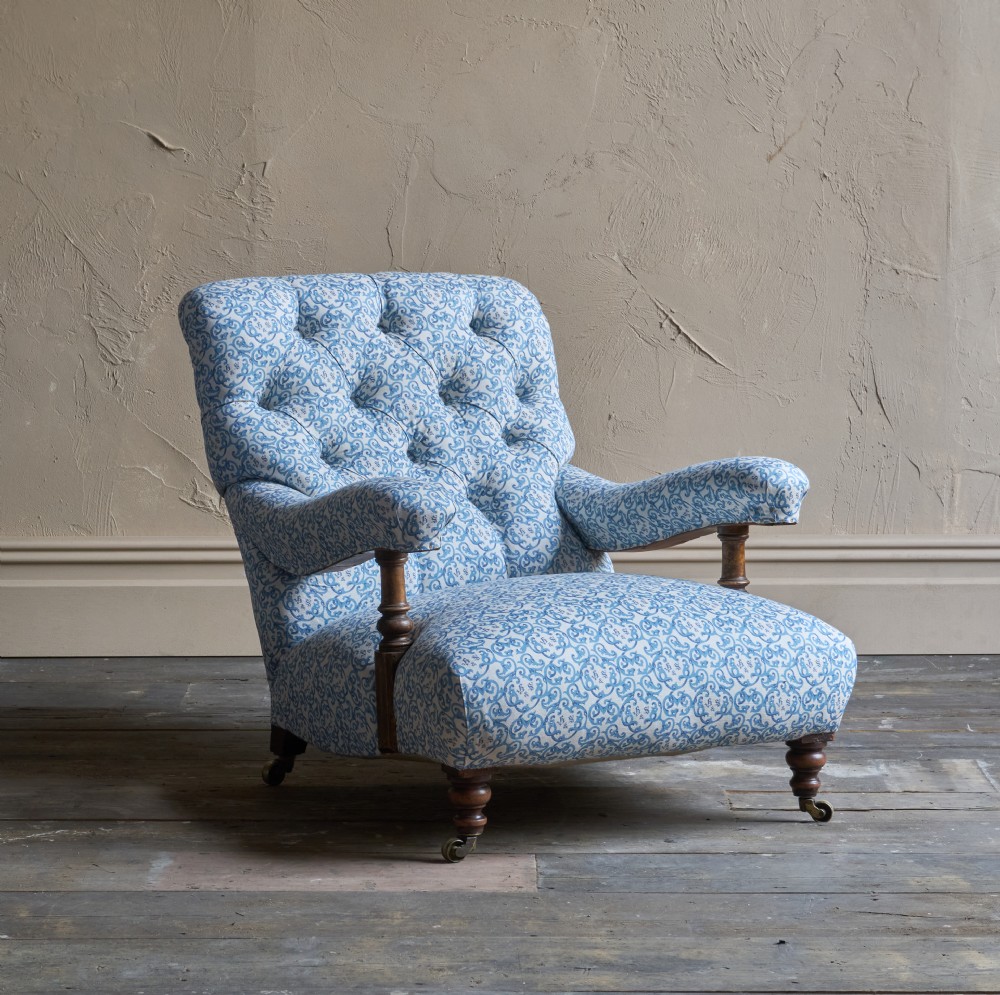 A low Victorian open armchair upholstered in a blue and white scroll pattern, featuring deep buttoning on the backrest, padded arms on turned wooden supports, and turned legs on castors.