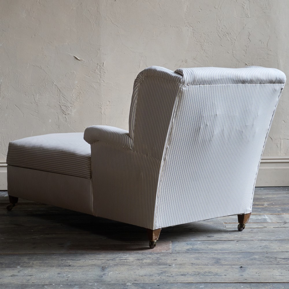 An upholstered antique chaise longue covered in off-white fabric with fine vertical stripes, featuring castors and a single high armrest. View VI