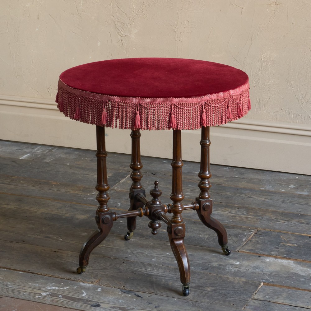 A round wooden side table with a deep red velvet top and fringed edge, supported by turned legs and castors.