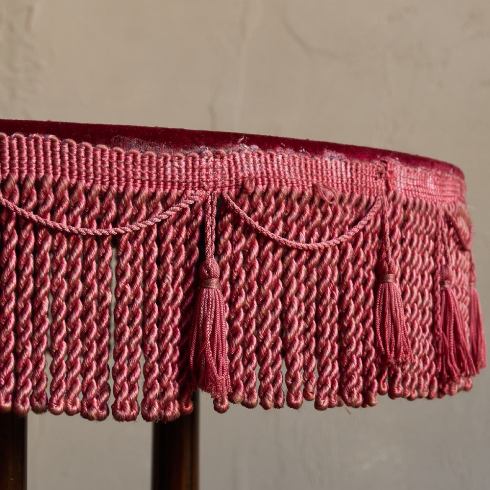 A round wooden side table with a deep red velvet top and fringed edge, supported by turned legs and castors. View III