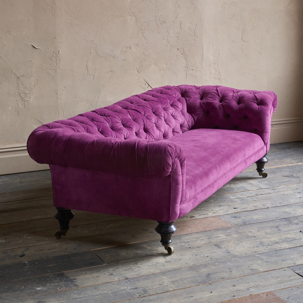 An antique velvet Chesterfield sofa featuring deep button tufting, rolled arms, and dark turned wooden legs with brass castors.