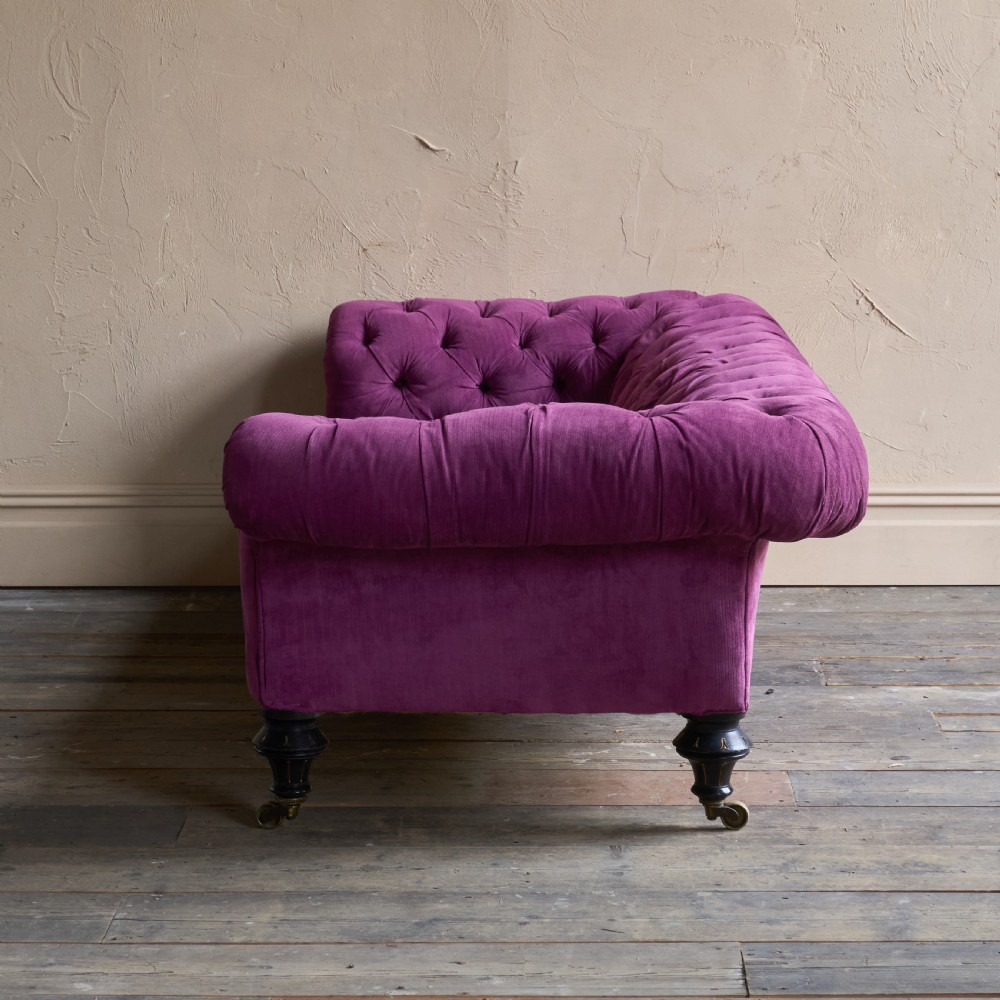 An antique velvet Chesterfield sofa featuring deep button tufting, rolled arms, and dark turned wooden legs with brass castors. View II