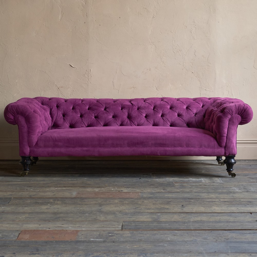 An antique velvet Chesterfield sofa featuring deep button tufting, rolled arms, and dark turned wooden legs with brass castors. View IV