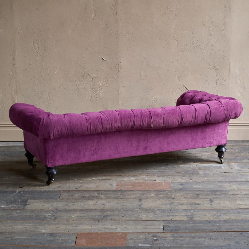 An antique velvet Chesterfield sofa featuring deep button tufting, rolled arms, and dark turned wooden legs with brass castors. View V
