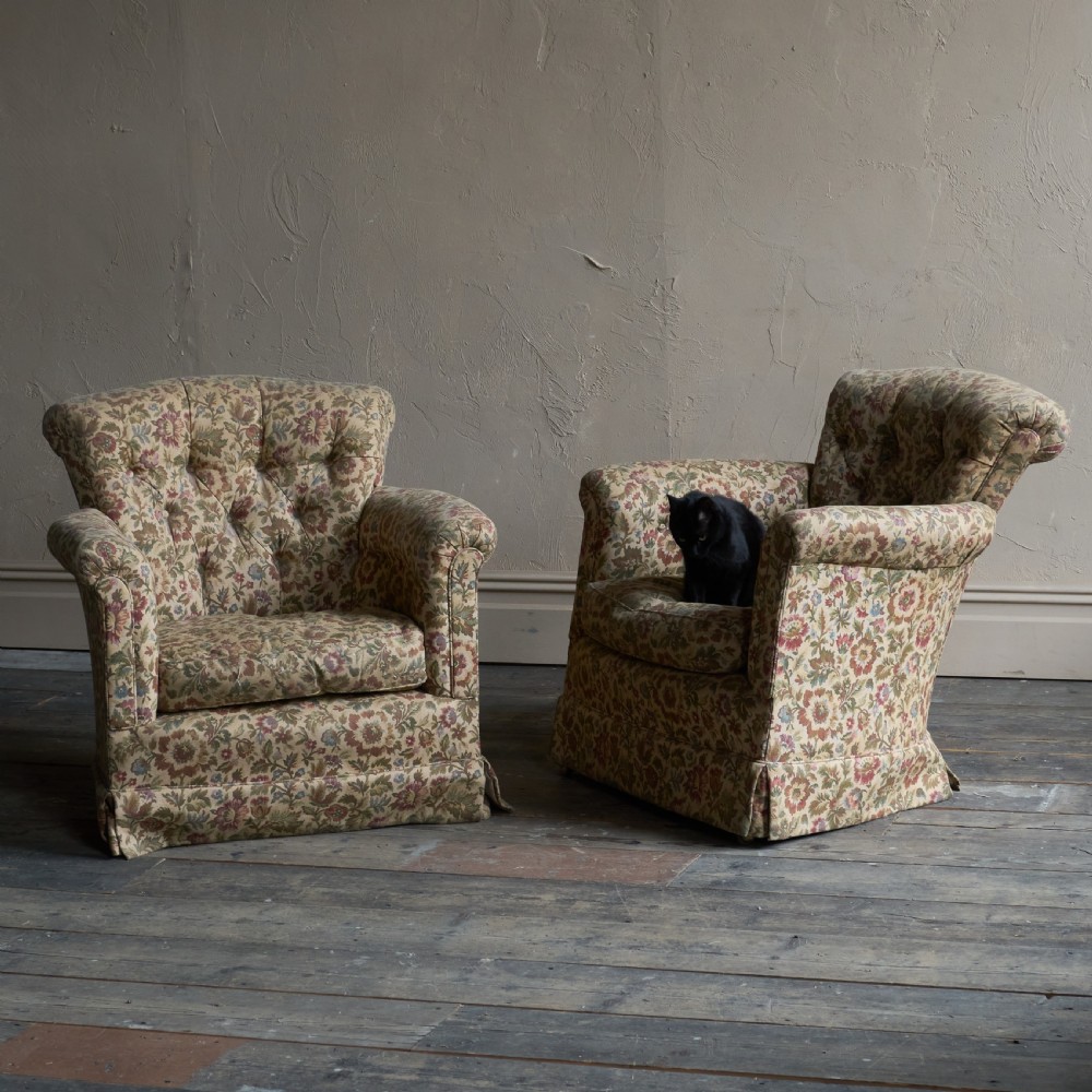 A pair of low-slung armchairs with deep-buttoned backrests, upholstered in a floral patterned fabric with rolled arms and skirted bases.