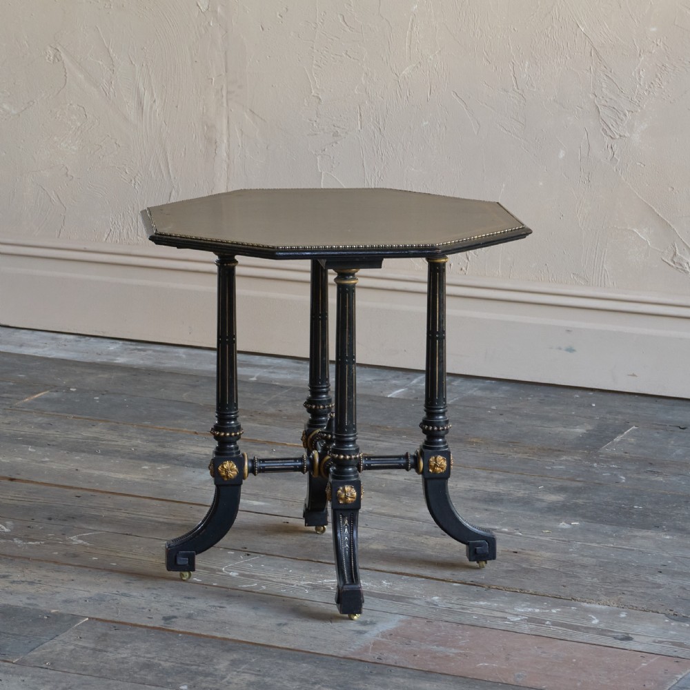 An octagonal ebonised side table featuring turned legs with gilt accents, central stretchers with carved rosettes, and brass castors.