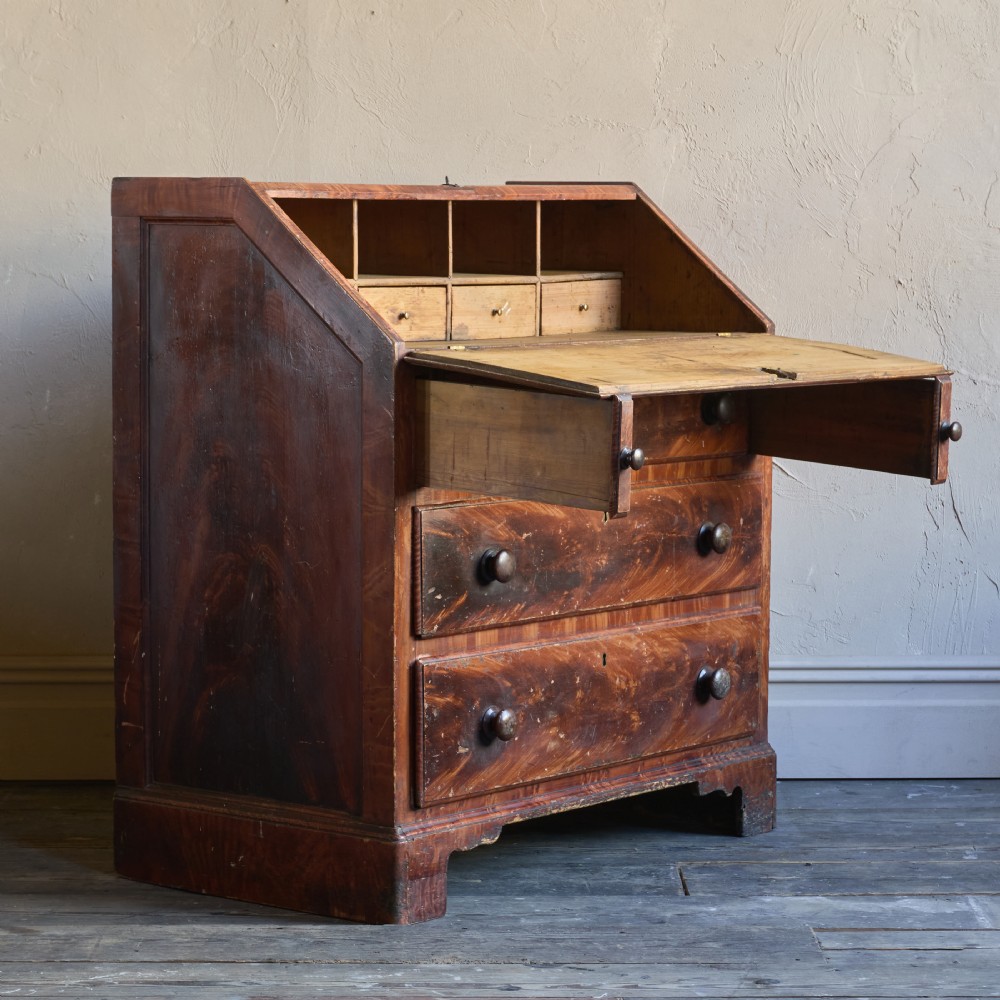 An antique wood bureau with a faux mahogany grain finish featuring a drop-down writing flap, internal pigeonholes, and three lower drawers with round turned handles. View IV