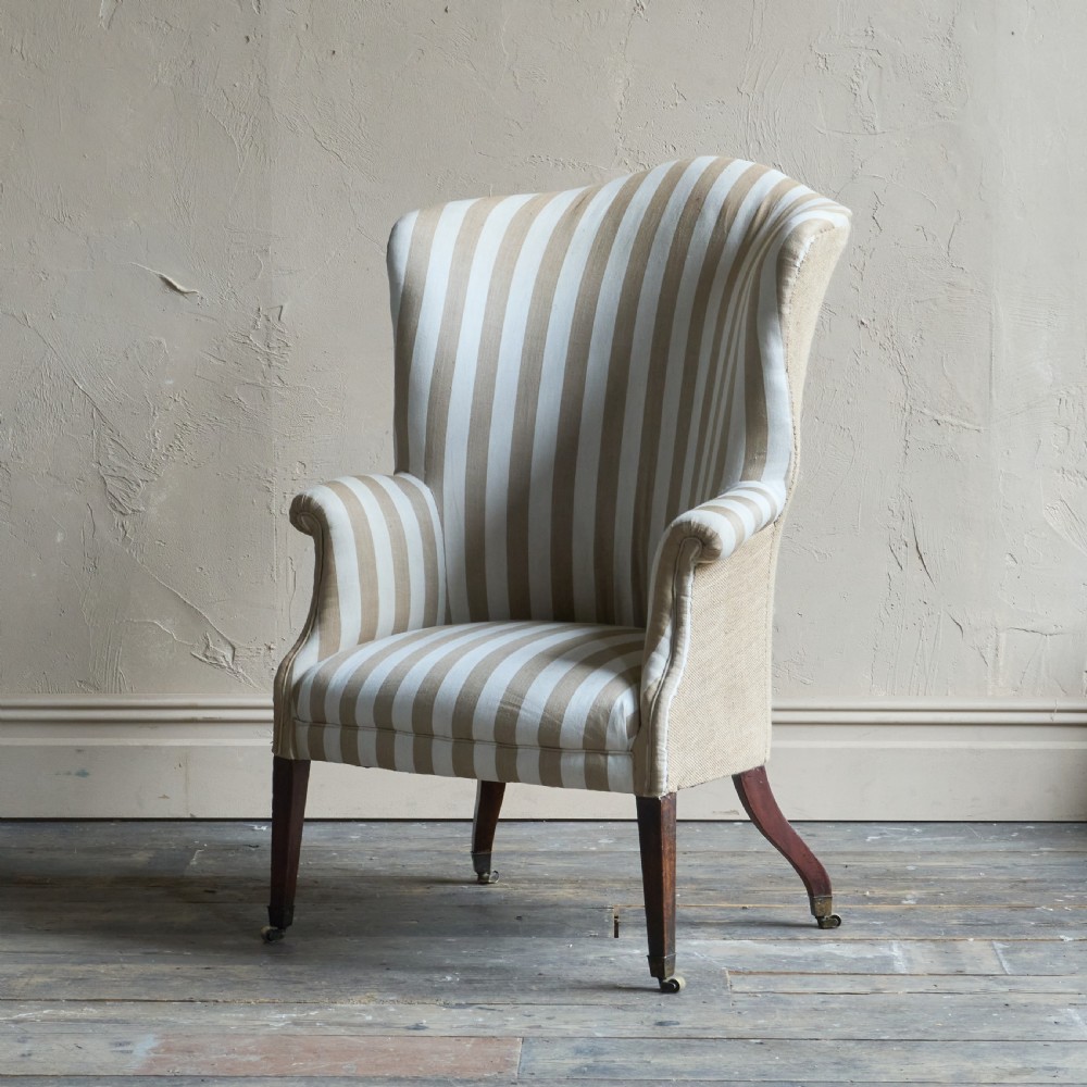An antique Georgian wingback armchair upholstered in neutral French ticking stripe fabric, set on dark wooden legs with castors.