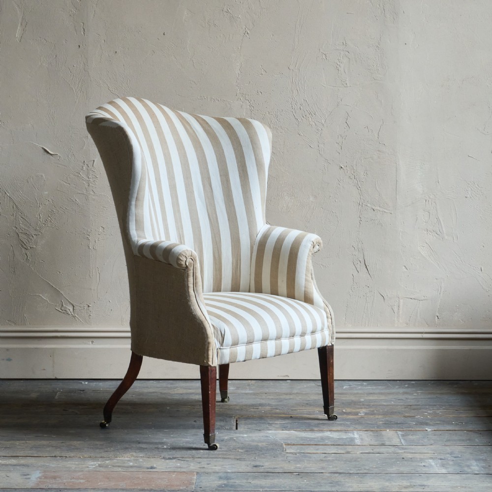 An antique Georgian wingback armchair upholstered in neutral French ticking stripe fabric, set on dark wooden legs with castors. View IV