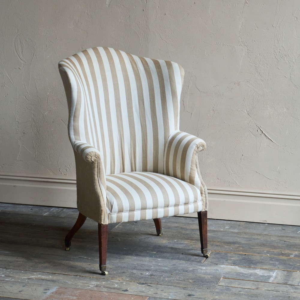 An antique Georgian wingback armchair upholstered in neutral French ticking stripe fabric, set on dark wooden legs with castors. View VI