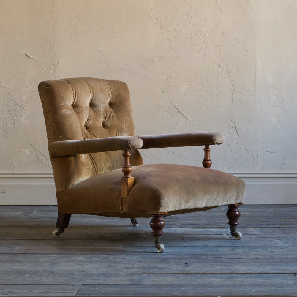 An antique open armchair upholstered in golden brown velvet featuring button-tufting, turned wooden front legs, and castors.