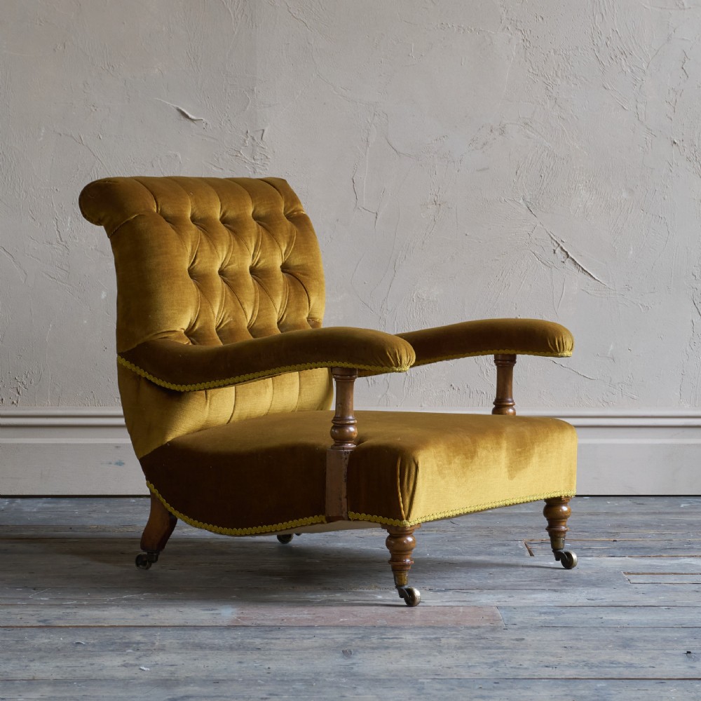 An antique open armchair upholstered in gold velvet featuring a button-backed scroll backrest, padded armrests on turned wooden supports, and turned front legs with brass castors.
