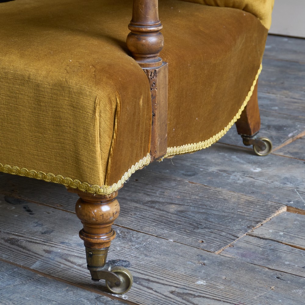 An antique open armchair upholstered in gold velvet featuring a button-backed scroll backrest, padded armrests on turned wooden supports, and turned front legs with brass castors. View VI