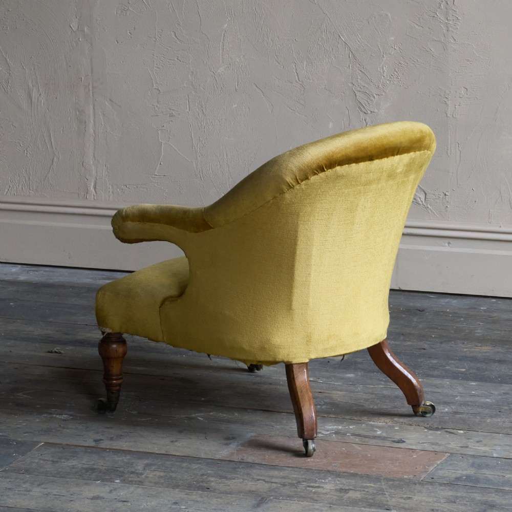 A Victorian low-slung armchair with a curved back, upholstered in mustard yellow velvet, featuring turned front legs with brass castors. View II