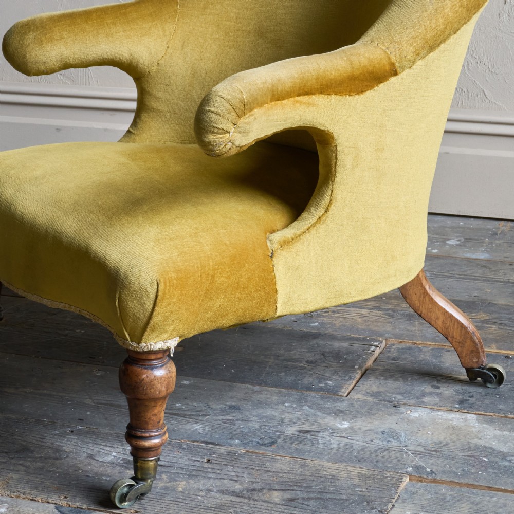 A Victorian low-slung armchair with a curved back, upholstered in mustard yellow velvet, featuring turned front legs with brass castors. View V