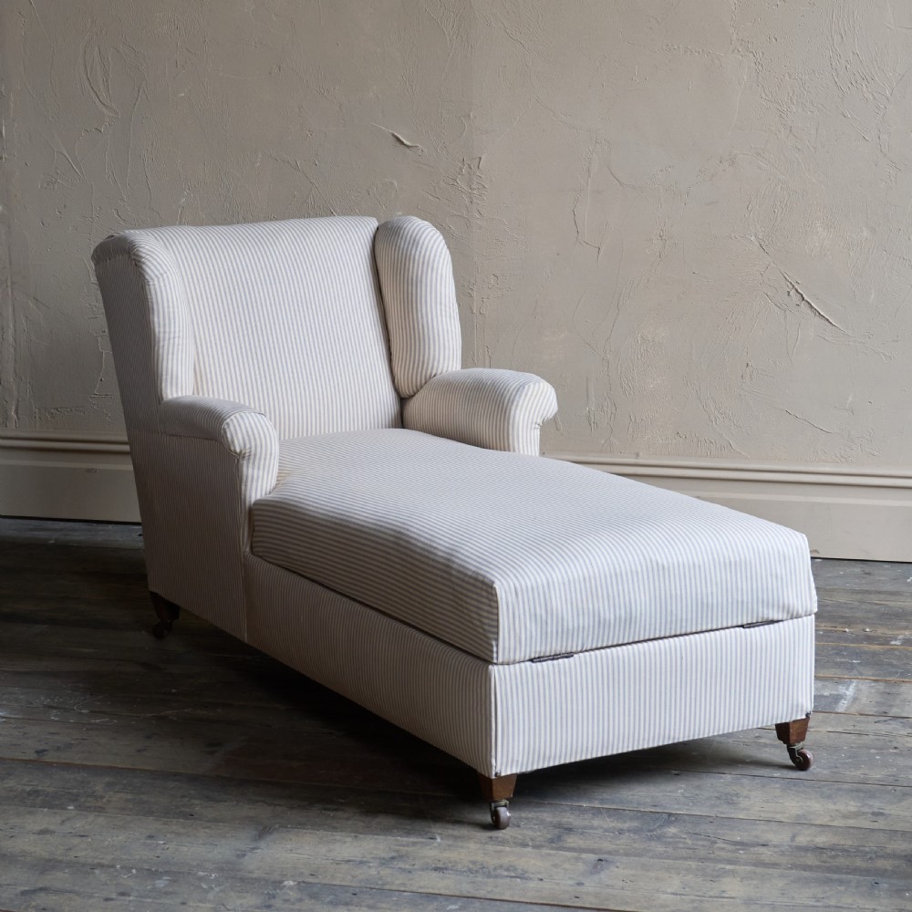 An antique upholstered chaise longue in a cream and beige ticking stripe fabric with a high winged backrest and integrated storage compartment.