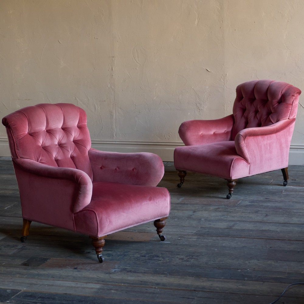 A pair of deep-seated Howard style armchairs upholstered in rose pink velvet featuring button-tufted backrests and turned wooden legs on castors.