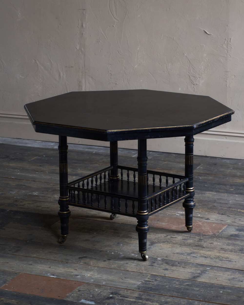 An antique ebonised octagonal centre table with gilded highlights, a lower galleried shelf, and turned legs on castors.
