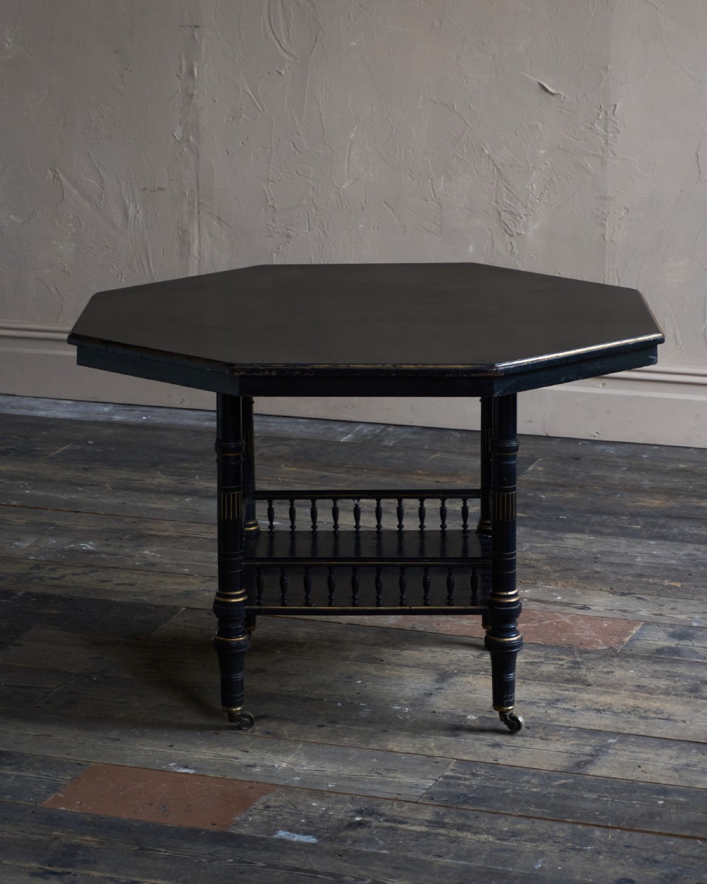 An antique ebonised octagonal centre table with gilded highlights, a lower galleried shelf, and turned legs on castors. View III