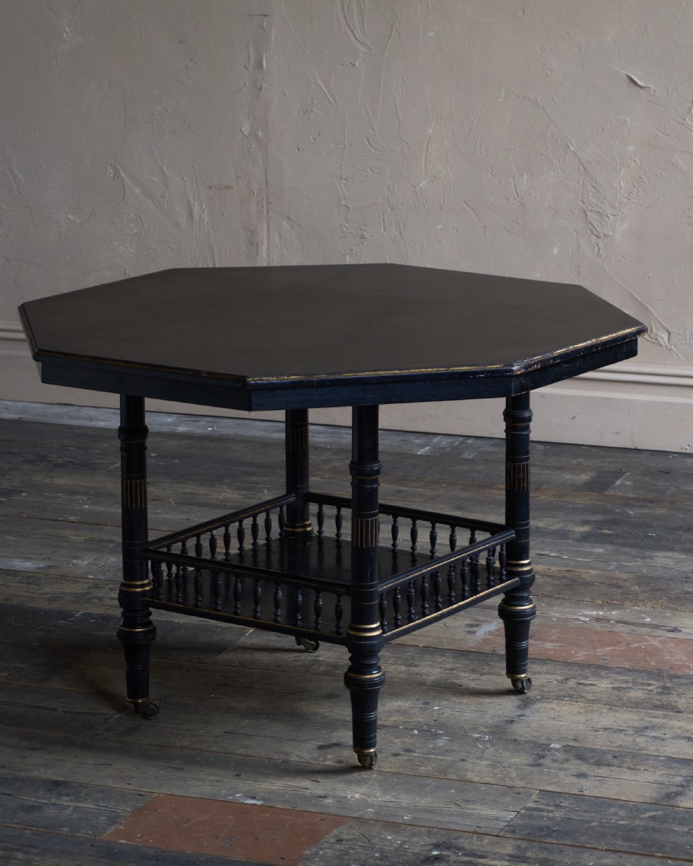 An antique ebonised octagonal centre table with gilded highlights, a lower galleried shelf, and turned legs on castors. View IV