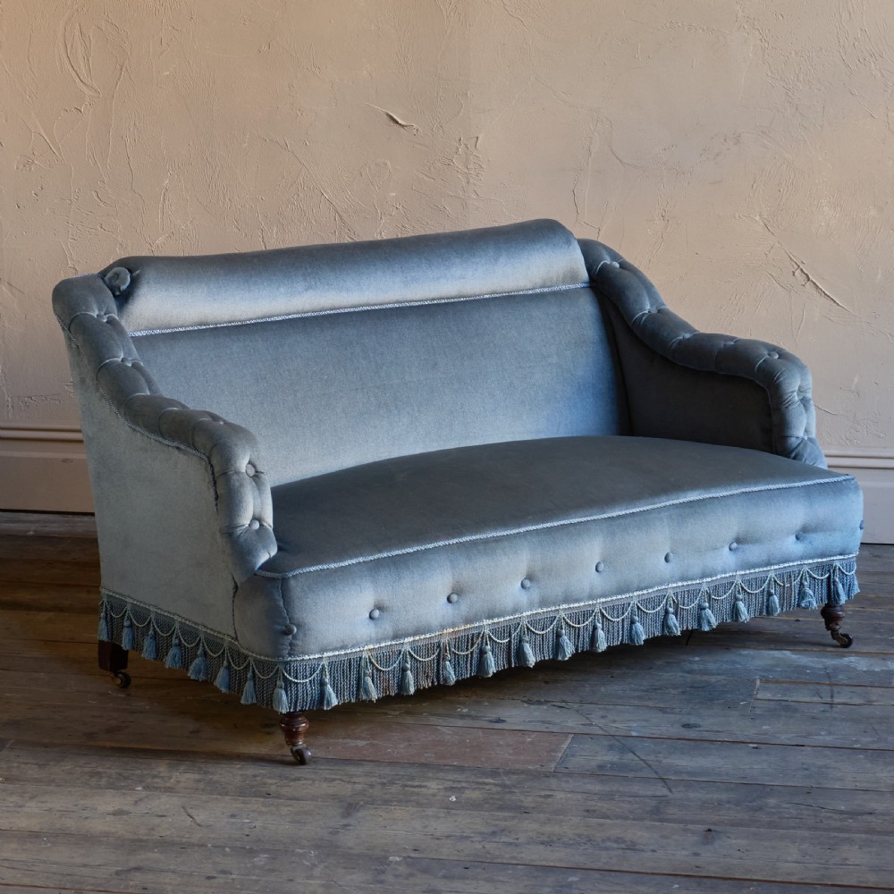 A Victorian bedroom settee upholstered in light blue velvet with buttoned details along the armrests and base, fitted with a tassel fringe skirt and turned wooden legs on castors.