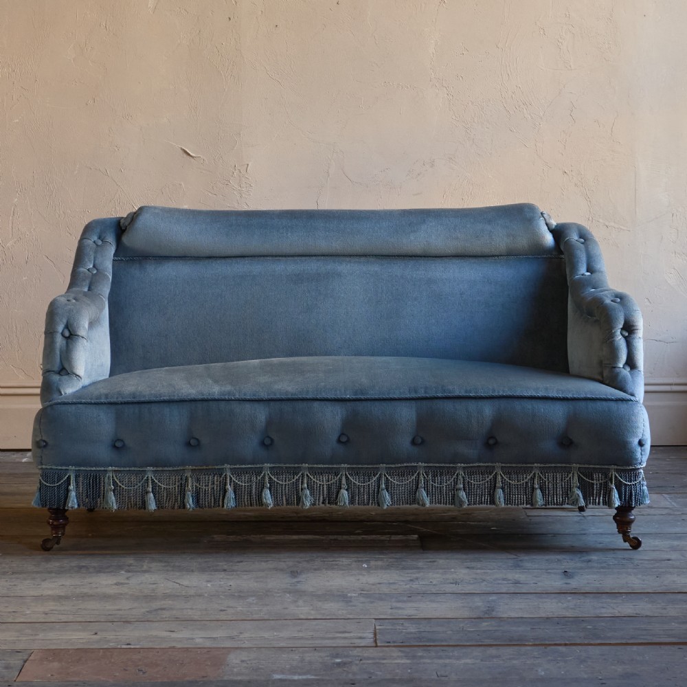 A Victorian bedroom settee upholstered in light blue velvet with buttoned details along the armrests and base, fitted with a tassel fringe skirt and turned wooden legs on castors. View II