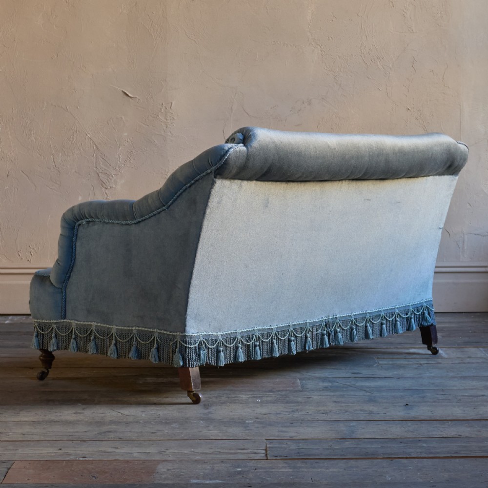 A Victorian bedroom settee upholstered in light blue velvet with buttoned details along the armrests and base, fitted with a tassel fringe skirt and turned wooden legs on castors. View V