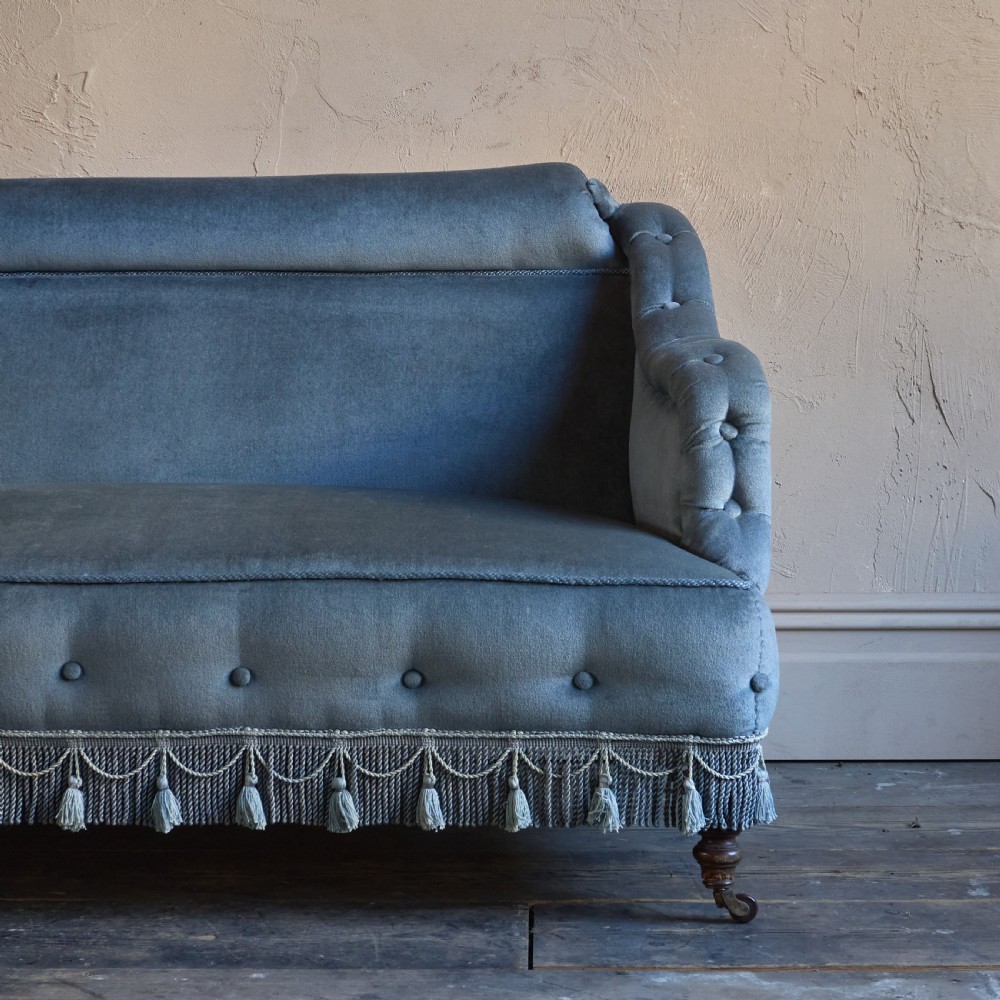 A Victorian bedroom settee upholstered in light blue velvet with buttoned details along the armrests and base, fitted with a tassel fringe skirt and turned wooden legs on castors. View VI