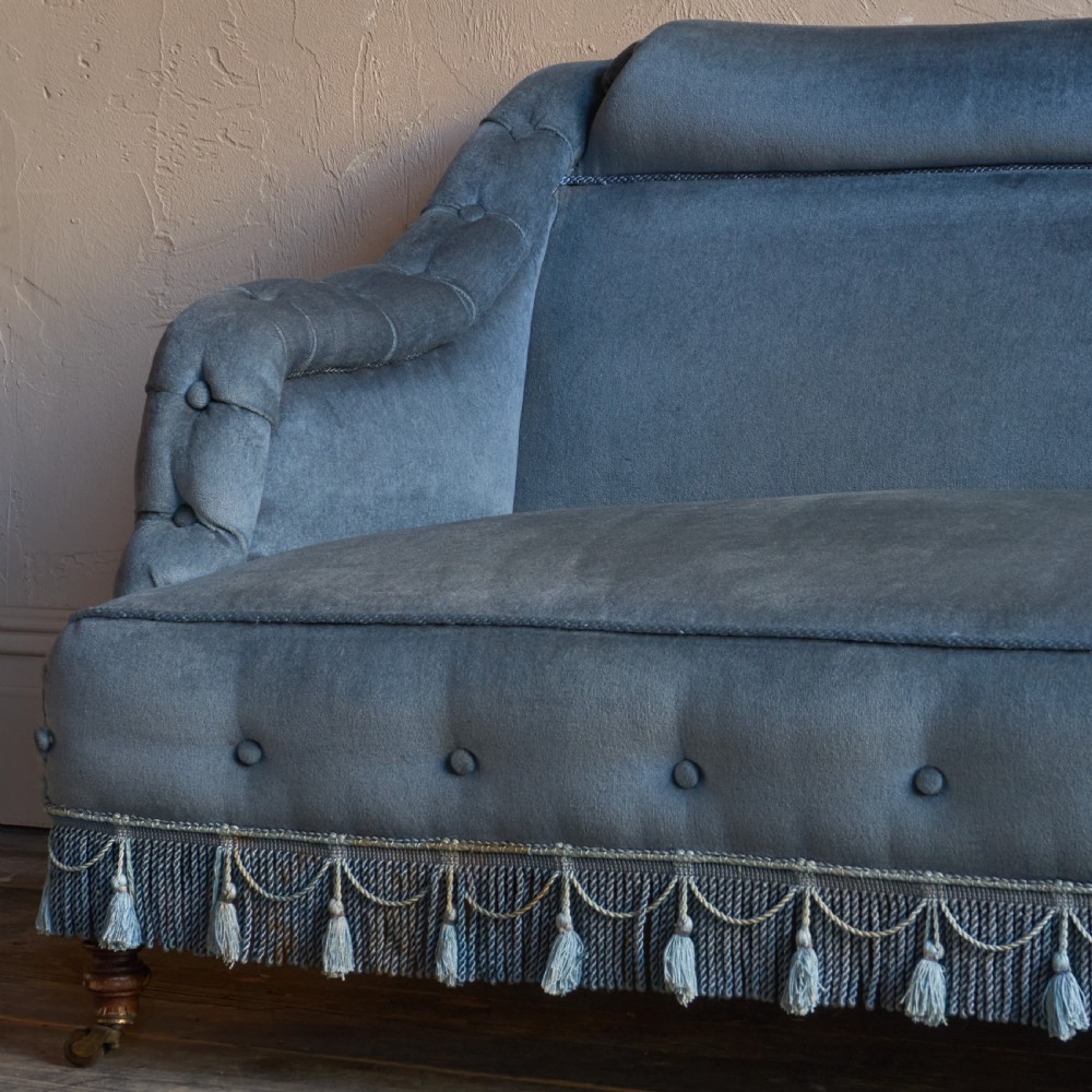 A Victorian bedroom settee upholstered in light blue velvet with buttoned details along the armrests and base, fitted with a tassel fringe skirt and turned wooden legs on castors. View VII