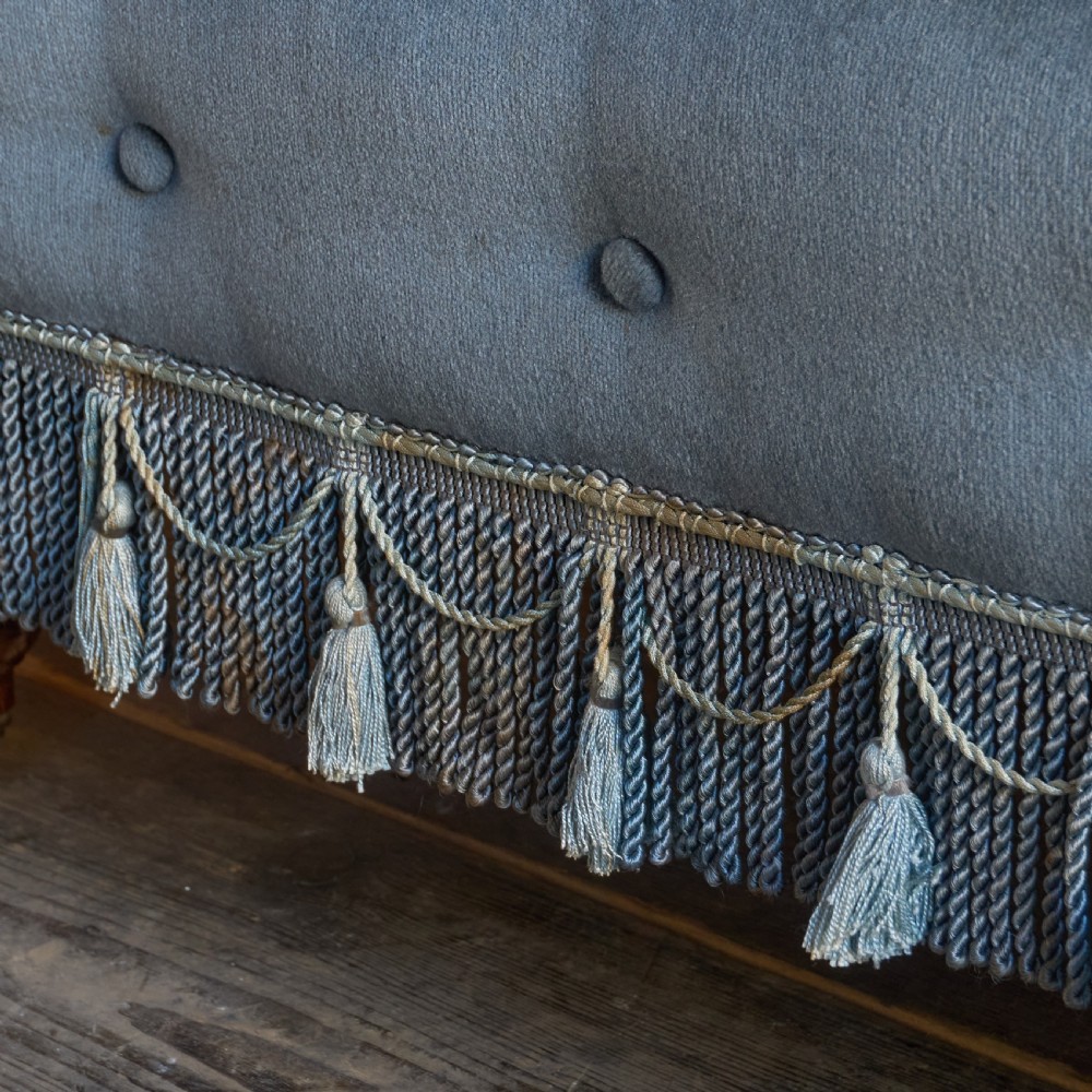 A Victorian bedroom settee upholstered in light blue velvet with buttoned details along the armrests and base, fitted with a tassel fringe skirt and turned wooden legs on castors. View VIII